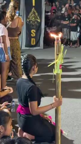 A medium, static shot shows a crowd of people in Bali, Indonesia watching a performance or event. In the foreground, a kneeling woman in a black shirt and pink sash holds a tall bamboo torch with a burning flame at its top. Green grass is tied to the pole. The woman has black hair in a long braid, and she looks at the torch in front of her. Other women are sitting or standing next to her, watching the scene. Behind them are two black banners with gold lettering that reads "STSD". In the background, a diverse crowd of people, some holding phones, watch the performance. The scene is outside at night, illuminated by streetlights.