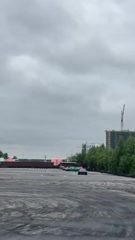 The video shows an outdoor asphalt track where cars are drifting. The sky is overcast. In the background, there are trees, buildings, and a construction crane. Several orange cones are placed on the track, possibly marking a course. Two cars are visible on the track, one black and one green, both in motion. The black car appears to be in the process of drifting, with smoke or dust kicked up from its tires. The green car is further back on the track.