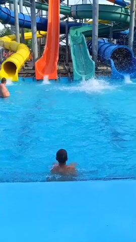 The video shows a water park with several colorful slides ending in a blue pool. In the foreground, a young boy, facing away from the camera, sits in the water at the edge of the pool. Further back, another boy swims in the pool. Three water slides are visible with water gushing out from their ends into the pool: an orange slide on the left, a green slide in the middle, and a blue slide on the right. To the far left, a yellow slide is also visible. A man is partially submerged in the water near the yellow slide. The water in the pool is clear and blue, with ripples and splashes created by the falling water and movement of people. The background shows the infrastructure of the water park, including metal supports for the slides. The overall scene depicts a recreational environment on a sunny day.