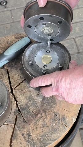 A close-up shot shows a person placing a metal dome-shaped object on top of a metal cylinder with a brass disc in the center. The objects are on top of a wooden stump with many cracks. To the left is a handle of a tool that has a grey handle. The background is blurred, showing a paved area.