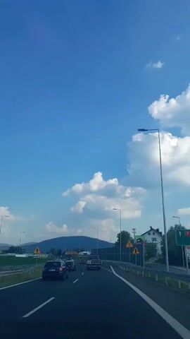 The video depicts a view from a vehicle traveling on a highway. The scene is set on a sunny day with a clear blue sky dotted with fluffy white clouds. The highway has multiple lanes, and several cars are visible, including a black sedan, a dark SUV, and a silver car. Streetlights line the right side of the road, and a guardrail separates the highway from the adjacent area.

In the background, rolling green hills or mountains are visible under the bright sky. There are signs along the highway, including triangular yellow warning signs with black symbols. One sign appears to indicate a sharp turn, while another shows a pedestrian crossing. A sign with a green background and white text, possibly indicating a route number, is also visible on the right.

To the right of the highway, a building with a white facade and a dark roof is present, with trees and foliage surrounding it. The overall atmosphere is one of travel and clear weather. The time of day appears to be daytime, likely late morning or afternoon, given the brightness of the sunlight and the shadows cast. The location is identified as Myślenice, Poland. No people are clearly discernible within the frame, and the focus is on the road and the surrounding landscape.