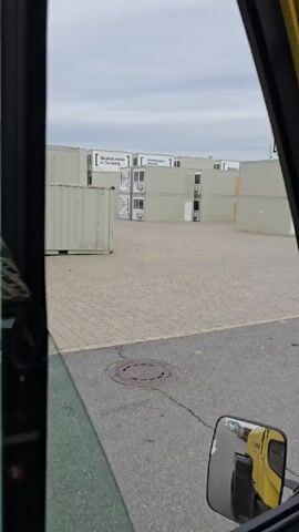 This is a first-person perspective video, likely from the operator's cabin of a forklift, looking out at a storage yard filled with modular buildings. The day appears overcast with a cloudy sky.

The main objects in the scene are rows of stacked, light-colored modular units, resembling shipping containers or prefabricated rooms. Some of these units have windows and what appear to be air conditioning units installed. They are arranged in orderly fashion across a paved lot, which has a textured, interlocking paver surface in the foreground and asphalt further back. There is a circular manhole cover visible on the paved area.

The forklift itself is partially visible, with its yellow frame, black hoses, and a red fire extinguisher mounted inside the cabin. A side mirror reflects part of the forklift's yellow body and the operator's seat.

Text is visible on some of the modular units. One prominently displays "Zeiträume" (meaning "time spaces" or "periods" in German) along with phrases like "Du steckst, was du bist" ("You are what you are") and "Flasche KIste" (which could mean "Bottle box" or be a brand name). Another unit has the text "Comfort made in Germany."

The activity appears to be the movement or organization of these modular units within a storage or logistics facility. The overall impression is of an industrial or commercial setting, likely a depot for prefabricated structures. The mood is neutral, typical of a work environment.