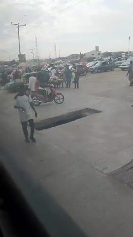 The video shows an outdoor scene with a paved surface, possibly a road or parking lot. Several people are visible, some walking, others standing. There are also numerous parked vehicles, including cars and a pink motorcycle. In the foreground, a man in a white shirt and dark pants is walking. A dark, rectangular hole is present in the paved surface near him. In the background, more people and vehicles are seen, suggesting a busy area. The sky is overcast.