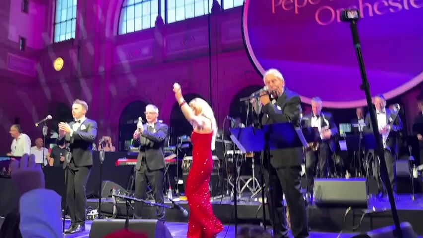 The video shows a live musical performance on a stage in a large, ornate building. In the foreground, a male singer in a tuxedo stands center stage, holding a microphone. To his left, a woman in a sparkling red dress dances with her back to the camera, facing the stage. To her left, two men in tuxedos are clapping and singing into microphones. Behind the performers, an orchestra is visible, with various musicians playing instruments such as saxophones and trombones. A large circular backdrop behind the orchestra displays the text "Pepe Lienhard Orchestra" in white lettering. The stage is lit with pink and purple lights. The audience is partially visible in the lower left corner of the frame, with some hands raised. A tripod-mounted camera is positioned to the right of the male singer.
