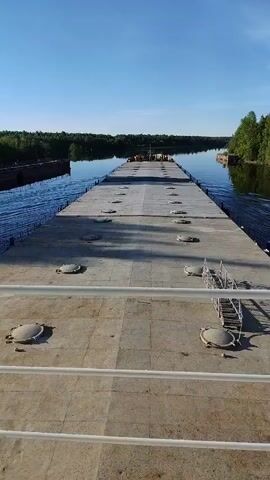 The video presents a forward-facing view from a vessel, likely a barge or ship, navigating a calm waterway. The foreground is dominated by a wide, flat deck, possibly the cargo hold or top of a large barge, featuring numerous evenly spaced circular hatch covers. A small, multi-tiered ladder or staircase structure is visible on the right side of the deck.

The vessel is moving through a narrow canal or river, which stretches towards the horizon. Both banks of the waterway are densely lined with lush green forest, reflecting in the placid water. The sky above is clear and blue, indicating fair weather and bright daylight, possibly morning or afternoon. No people are visible in the frame. The overall scene suggests peaceful transit through a natural, forested landscape in the area of Annensky Most, Russia. There is no visible text.