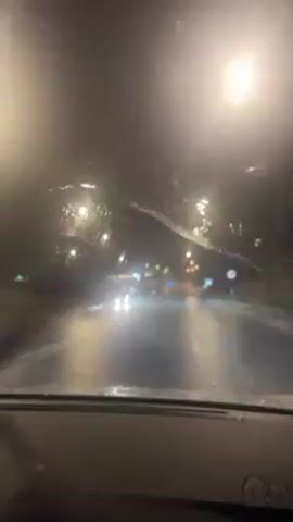 The video depicts a night-time car ride in the rain. The view is from inside the car, looking out through the windshield. Raindrops are visible on the glass, and streetlights and other cars' headlights illuminate the wet road ahead. The road appears to be paved, and the car is moving forward.