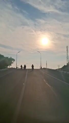 The video shows a slightly shaky, first-person perspective of a road, likely a bridge, on a sunny day. The sun is high in a bright blue sky with wispy white clouds. Several motorcycles, silhouetted against the sun, are traveling in the same direction up the road. Streetlights line the sides of the road, and there are trees and buildings visible in the distance. The camera appears to be mounted on a moving vehicle, possibly another motorcycle or a car, judging by the slight movement and vibration. The overall impression is that of a peaceful, early morning or late afternoon journey.
