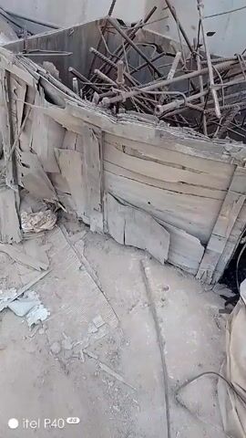 The video shows the interior of a cluttered, damaged building, followed by an exterior scene of a construction or industrial area. 

Inside, there's debris consisting of broken wood, metal rods, white cloth or tarpaulin, cardboard, and piping. The interior walls appear to be light-colored. The structure is damaged and appears to be in a state of disrepair.

Outside, there are stacks of metal sheets, pipes, rods, a rusty barrel, and various pieces of trash scattered on a dirt surface. In the background, there are green shipping containers.
