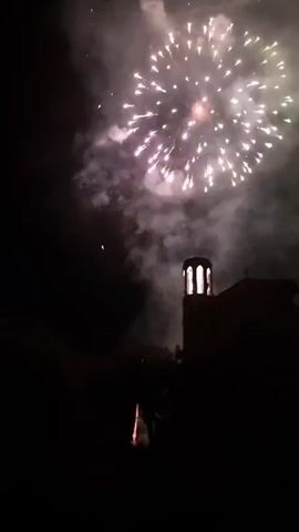 This media file captures a nighttime fireworks display. The main subjects are the bursts of light from the fireworks, illuminating the dark sky. In the foreground, a silhouetted tower with arched windows is visible, likely part of a building in Sant Joan Les Fonts, Spain. Trees are also silhouetted against the night sky.

The event taking place is a fireworks show, with multiple explosions of light and trailing sparks. The time of day is clearly night, indicated by the darkness and the presence of fireworks. There is no visible text in the media file. The mood evoked is celebratory and awe-inspiring, typical of a fireworks display. The location cues suggest a public event or celebration in a town or city setting.