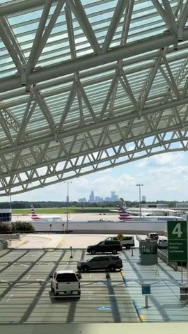 The video shows a view from an airport terminal looking out at the tarmac. A large, modern airport roof structure with white metal beams and a glass ceiling is in the foreground. Below the roof, several cars are parked in a lot, casting shadows on the ground. Beyond the parking lot, two American Airlines airplanes are visible on the tarmac, with one parked and the other possibly taxiing. In the distance, a city skyline with skyscrapers can be seen against a blue sky with scattered clouds. A green signpost labeled "4 American Etihad Lufthansa" is also visible near the tarmac. The overall scene depicts a busy airport environment with a view of the city in the background.