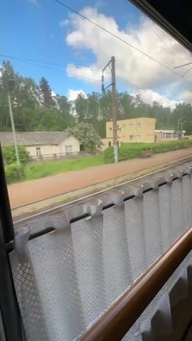 The video shows a view from inside a train car, looking out the window. The train is moving past a train station. The station has a yellow building, a platform, a bench, and a small outbuilding. There are trees and a blue sky with clouds visible in the background. The train car has a white lace curtain and a wooden handrail. The inside of the train car is visible, showing the corridor with doors to passenger compartments.
