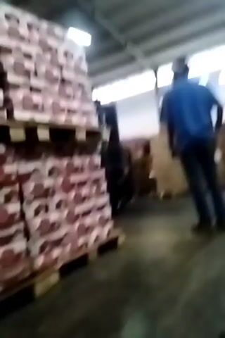 A man wearing a blue shirt and dark pants stands in a warehouse, facing away from the camera. He is looking towards stacks of boxes on pallets. A forklift is visible in the background, between the man and the boxes. The warehouse appears to be lit by overhead lighting.