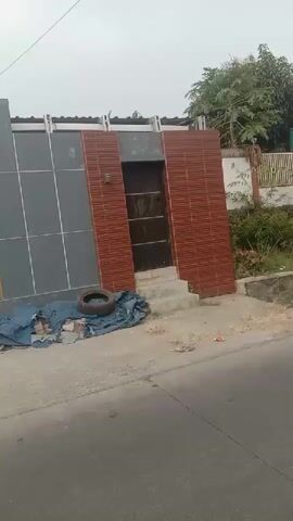 The video shows a street scene, likely in a residential area or near a school. The first few seconds show a house with a modern gray facade and a reddish-brown tiled section. In front of the house, there is a pile of dark blue tarpaulin with a tire resting on top. The camera then pans to the left, revealing a street with several children, some wearing school uniforms with white shirts and red shorts or skirts. Some children are sitting by the side of the road, while others are walking or running. There are buildings on the right side of the street, one with shops displaying banners. The video continues to show the children interacting on the street, with a motorbike mirror visible in the foreground for a few seconds. The final shot shows one of the children running past a large wooden gate. The environment appears to be outdoors with an overcast sky.