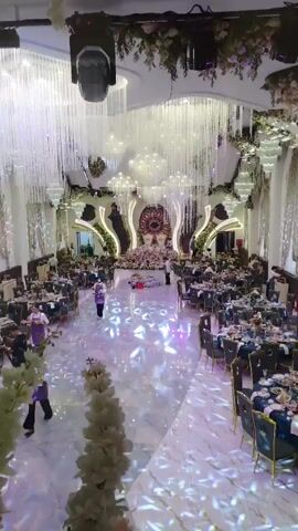 The video shows a high-angle view of a large, elaborately decorated event hall, likely set up for a wedding reception. Rows of round tables, draped with dark blue cloths and adorned with floral centerpieces and place settings, fill the sides of the hall. In the center, a wide aisle leads to a grand stage area, decorated with ornate golden frames, floral arrangements, and a large circular backdrop featuring intricate patterns. Numerous strings of sparkling crystals hang from the ceiling, creating a dazzling effect, along with various light fixtures and chandeliers. Several individuals are visible throughout the space. Two people in purple uniforms appear to be staff, one walking down the aisle with a tray, and another near a table. Another person in dark clothing stands by a camera on the left side of the aisle, suggesting a photographer or videographer. More figures are seen seated at the tables or standing around them, dressed in formal attire. The floor is highly reflective, with patterned lights projected onto it, enhancing the opulent atmosphere.