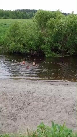 The video shows three individuals in a river. The person in the center is standing up in the water, with their arms raised in a peace sign gesture. Two other individuals are visible in the water near the central figure. The river has dark water and is surrounded by lush green trees and foliage. The foreground of the video features a sandy area. The weather appears to be overcast.
