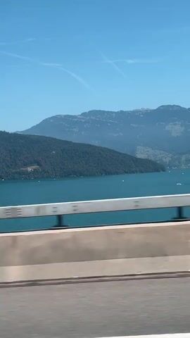 The video shows a wide shot from a moving vehicle of a serene landscape under a clear blue sky. A large, calm lake with scattered sailboats dominates the middle ground, bordered by lush green hills and distant mountains. A highway barrier runs horizontally across the foreground, with blurred greenery on either side, indicating the vehicle's movement. The overall impression is one of a peaceful journey through a picturesque natural setting.