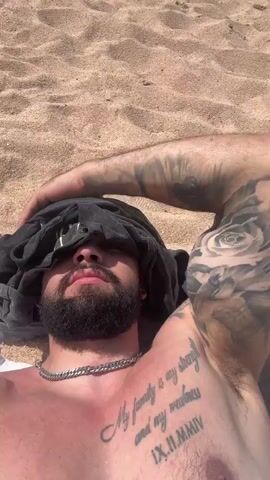 The video shows a close-up, overhead shot of a man lying on a sandy beach. He is wearing a silver chain necklace and has tattoos on his arm and chest. A black t-shirt is draped over his face, obscuring his eyes. The man appears to be relaxing or sleeping. The background is composed of sand, suggesting an outdoor beach setting.