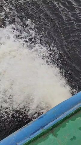 The video captures a close-up, high-angle view of water being churned up by a moving vessel. The water is dark and appears agitated, with a significant amount of white foam and spray being generated. The edge of the vessel is visible in the foreground, with a blue railing and a green surface. The movement suggests the boat is traveling at a moderate to fast speed. No people are visible in the frame.