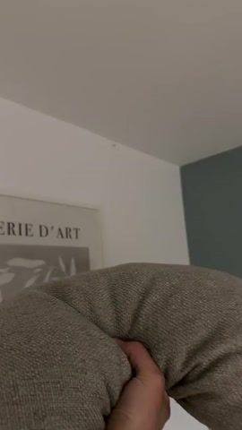 A person's hand is visible on the right side of the frame, holding a textured, beige pillow. The pillow is positioned in the foreground, obscuring a significant portion of the scene. In the background, a white wall is visible, with a framed picture on the left side. The picture has text that reads "IE D'ART" and appears to be a minimalist art print. The top of the frame shows a white ceiling and a corner where the wall meets another wall painted a muted green. The camera appears to be held at a low angle, looking upwards. The pillow is then thrown upwards and out of frame, disappearing from view.