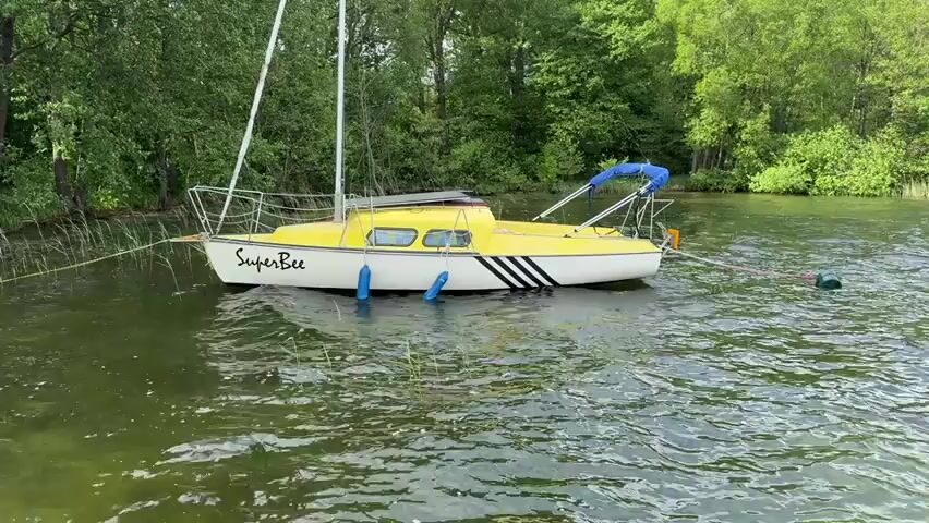 A yellow and white sailboat, named "SuperBee", is moored in a lake. The boat has a tall white mast, a yellow cabin, and three black stripes on the side.  The water is rippling. The shoreline is visible in the background, covered in green trees.  The boat is tied with a yellow rope to the shore and a green buoy is tied to the boat in the back. The camera is stable.