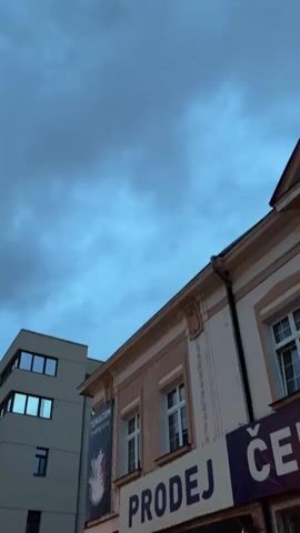 The video begins with a low-angle shot of buildings against a cloudy, blue sky. The camera pans slightly to the right, revealing more of the architecture, including windows, awnings, and a banner that reads "PRODEJ." There are glimpses of other buildings, some appearing modern and others older. As the video progresses, the camera continues to move, showing more of the streetscape, including streetlights and what appears to be construction fencing. The sky remains cloudy, with the light suggesting it is either early morning or late evening. No people are clearly visible in the video.
