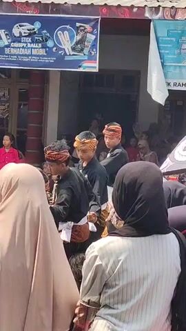 A group of young people in traditional clothing are performing some kind of martial art demonstration. They are wearing black shirts with colorful sashes and headbands. They are in front of a building with a banner that advertises a product called "STICK CRISPY BALLS". A crowd of people are watching the performance. The event appears to be taking place outdoors on a sunny day.