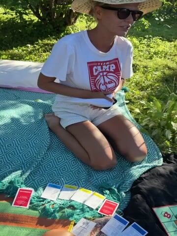 A group of people are playing Monopoly outside on a blanket. The Monopoly board is set up in the center, with cards and money scattered around it. The sun is shining and the grass is green. One person is wearing a white t-shirt and shorts. Another person is wearing pink Crocs. The video is taken from a high angle, looking down at the game.