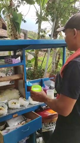 The video shows a man wearing a black t-shirt, a red towel around his neck, and a black baseball cap, standing behind a blue food cart attached to a motorcycle. The food cart has several compartments displaying food items, including what appears to be rice and toppings in plastic bags, and bottles of condiments. The man is holding a green cup and a spoon, and is in the process of preparing food, seemingly adding a condiment to a dish. The background suggests an outdoor setting with trees and foliage. The man appears to be interacting with the food cart, possibly serving a customer, although no other person is visible in the frame.