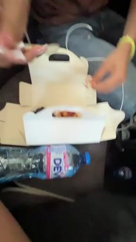 The video shows the interior of a bus. The initial shot focuses on a woman with long, dark hair and wearing a maroon top and ripped jeans. She is seated and opening a white cardboard box containing small containers of berries and granola. A bottle of water is visible nearby. The camera angle then shifts to a young man with short brown hair wearing a black t-shirt. He looks at the camera and briefly makes a hand gesture. The video ends with a close-up shot of a leg covered in ripped jeans. The setting appears to be cramped, with various items scattered around the seats.