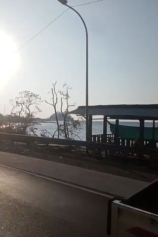 This video shows a truck driving on a road. The truck is white with a yellow bed. The truck has a flag attached to it. There are trees and a body of water in the background. The sun is shining.