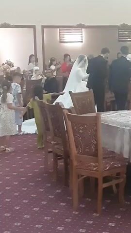The video shows a wedding reception. A bride in a white wedding dress with a veil is walking with a groom in a black suit. They are surrounded by guests. Children are also present. Some guests are seated at tables covered with tablecloths. The room is decorated with floral arrangements and has a patterned carpet. The lighting appears to be artificial.