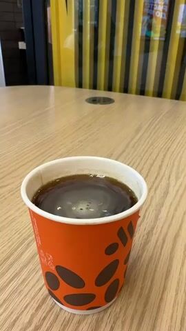 A close-up, eye-level shot focuses on a disposable orange coffee cup filled with dark coffee. The cup, adorned with brown oval shapes resembling coffee beans, rests on a light wood-grain table. The surface of the coffee shows subtle ripples, indicating it might be hot or recently stirred. In the background, a yellow and black striped pattern is visible, possibly part of a restaurant's decor. The lighting is bright, casting a slight shadow to the right of the cup. No people are visible in the frame.