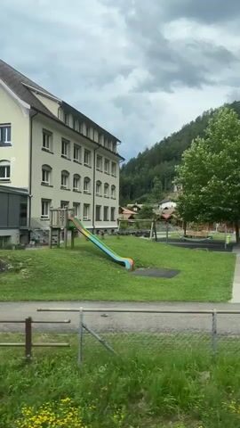 The video captures a view of a schoolyard and surrounding buildings, likely from a moving vehicle. On the left, a multi-story building with numerous windows is visible, with a colorful slide in the foreground. The schoolyard features a grassy area, a paved path, a swing set, and what appear to be picnic tables. In the background, a wooden building with solar panels on its roof is present, situated against a backdrop of lush green trees and a cloudy sky. A fence with red and white caution tape runs across the scene, suggesting some area is cordoned off. The overall impression is that of a tranquil, perhaps rural, educational setting.