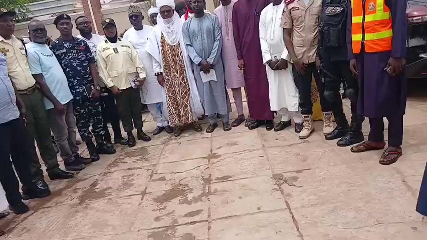 A group of approximately 20 men, dressed in various uniforms and traditional attire, are posing for a photograph outdoors. The group includes individuals in what appear to be security or law enforcement uniforms, as well as men in traditional Nigerian clothing. Some of the uniforms are khaki, some are dark blue with camouflage patterns, and one man is wearing a high-visibility orange vest. The traditional attire includes long flowing robes and head coverings, with one man wearing a white turban.

The individuals are standing on a paved surface that appears to be outdoors, with some fallen leaves or debris on the ground. Behind them, there is a white wall, a building with a brown tiled roof, and some greenery. The lighting suggests it is daytime, likely late morning or early afternoon, with overcast conditions. The expressions on the faces of the people are neutral, as is common for posed photographs. The overall scene suggests a formal gathering or official visit, possibly in a public or administrative building in Sabon Gari North, Nigeria. There is no visible text in the image.
