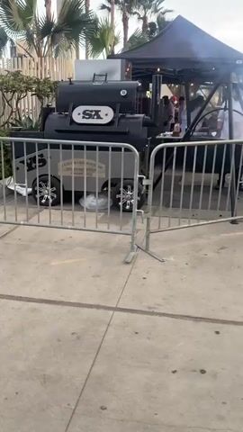 The video shows a barbecue smoker on wheels that appears to be on a paved surface. The smoker is black and has a logo that says "SX". There is a fence in front of the smoker, and behind it, there is a tent and some people who appear to be working. There are also some palm trees and bushes in the background. The video is shaky and the lighting is dim.