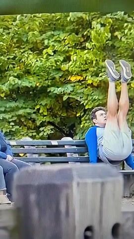 A young man is performing a handstand on a park bench. Another young woman is reaching towards him. They both fall off the bench and onto the ground. Two young women are sitting on a park bench laughing.