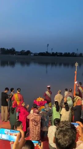The video shows a large group of people gathered near a body of water at dusk.  Several individuals in the foreground are holding up smartphones, seemingly recording the event.  In the middle ground, a group of people are participating in a ritual.  Two individuals, dressed in what appear to be religious or ceremonial attire with red and yellow sashes, are holding lit torches or lamps.  One of these individuals is standing near a small, decorated altar-like structure on which there are several lit diyas (oil lamps).  Another person in ceremonial attire is holding a trident-like object with a lit lamp at the top.  The rest of the gathered crowd, composed of men, women, and children, are facing the water and the ceremony, some appearing to be observing with interest.  The water's surface reflects the dim light.  In the background, the sky is a deep blue, transitioning into twilight.  Distant lights and structures are visible across the water, suggesting an urban or semi-urban environment.  The overall atmosphere suggests a religious or cultural ceremony taking place by the water's edge.