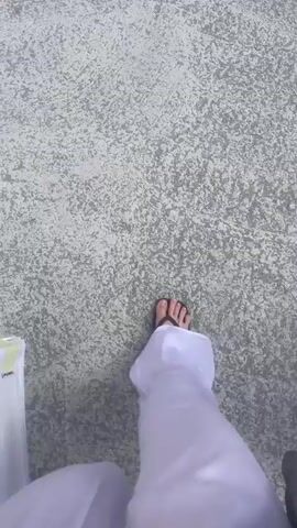The video begins with a first-person perspective of someone walking. Their feet, clad in sandals, and the hem of white trousers are visible. The ground appears to be a light-colored, textured surface, possibly concrete or asphalt, with a dappled grey and white pattern. The camera then pans slightly upwards and moves forward, revealing more of the path. The path is made of the same light-colored material and appears to be leading towards a tunnel. The tunnel entrance is arched and made of concrete, with some graffiti visible on its sides. The surrounding environment is dry and arid, with rocky hillsides and sparse vegetation, including some dry grass and a small tree on the left. The lighting suggests it is daytime, with sunlight illuminating the scene. The video continues to show the path leading towards the tunnel, with the camera moving at a walking pace.