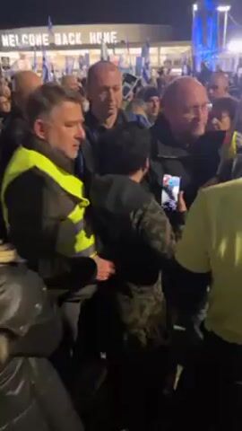 The video shows a chaotic scene outdoors at night, with a large crowd of people gathered. Many individuals are wearing yellow vests. Some people are holding flags. One person is holding up a phone, possibly recording. The crowd is densely packed, with people jostling each other. A building with illuminated windows and a sign reading "WELCOME BACK HOME" is visible in the background. The lighting is mixed, with streetlights and building lights illuminating the scene.