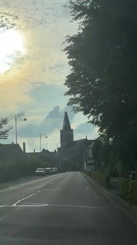 A first-person perspective video shows a slow approach down a paved road towards a church. The sky is partly cloudy with the sun visible on the left side of the frame, casting a light on the clouds. Several cars are parked on the left side of the road. The church is tall and made of stone with a prominent spire. Trees and hedges line the right side of the road. The overall impression is a calm, almost serene scene in a small town.