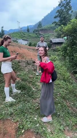Three individuals are captured in a daytime outdoor scene in Nậm Dịch Commune, Vietnam. The setting is a rural, hilly landscape with lush green vegetation, red-brown earth, and simple houses with corrugated iron roofs scattered on the slopes. Power lines are visible in the distance against an overcast sky.

In the foreground, a woman with curly brown hair, wearing a green crop top, white shorts, white knee-high socks, and white sneakers, is engrossed in her smartphone. To her right, a woman with long blonde hair, dressed in a black crop top, a red jacket draped over her shoulders, a long grey skirt, and light-colored slide sandals with pink socks, is actively talking and gesturing with her hands, holding a small clear bottle. She has visible tattoos on her left arm and is smiling animatedly. In the background, a man with a beard and a cap, wearing a grey t-shirt and green shorts, stands holding a camera, observing the other two. The ground is a mix of dirt and green grass. No visible text is discernible.