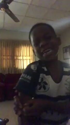 A child is standing indoors in what appears to be a living room. He is wearing a short-sleeved shirt with a white front and patterned dark sleeves. He initially has his arms crossed. The child looks upward and laughs loudly with his mouth open wide. He then looks directly at the camera, continuing to laugh. Subsequently, he lowers his gaze. The room contains a ceiling fan, curtains partially covering a window, framed pictures on the wall, and a burgundy-colored couch.