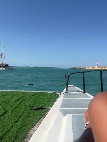 A static, full shot captures the view from a boat sailing on a body of water under a clear, blue sky. Another boat, a sailboat, is visible in the distance, filled with people. To the right, a small lighthouse with red and white stripes stands on a rock formation. In the foreground, the deck of the boat is visible, covered in artificial turf. A person's bare legs and feet, adorned with a shell anklet, are resting on the deck. The water is choppy, creating small waves. The overall scene suggests a bright, sunny day on the water.
