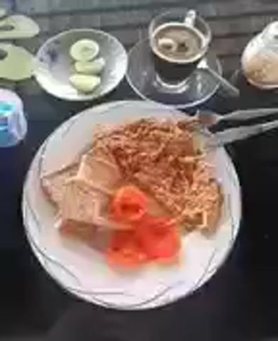 The video shows a table set with a meal, coffee, and lemon slices. The plate contains what appears to be breaded food covered with shredded topping, accompanied by tomato slices. A man sits at the table, holding a drink. Behind him, a lush green landscape stretches into the distance, partially obscured by mist or fog. The scene seems to be captured from a covered outdoor seating area.