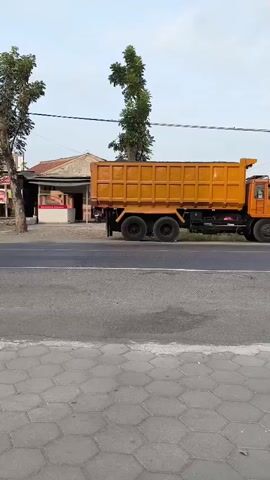 A dump truck and a scooter are on a road. The dump truck is orange and has a large bed. The scooter is white and has two people on it. The scooter is in front of the dump truck. The road is paved. There are trees and buildings in the background. The sky is cloudy.