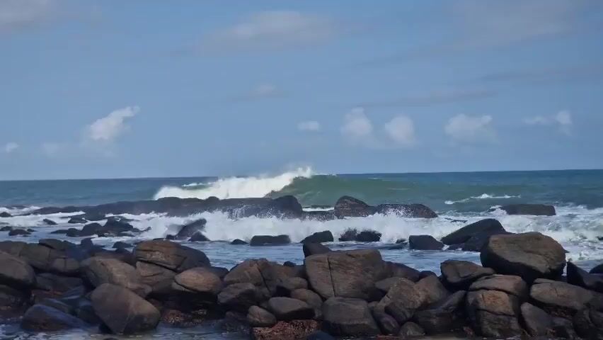 The scene depicts a rocky beach on a partially cloudy day. Large, dark brown rocks form the foreground, while the ocean occupies the middle ground, with waves crashing against the rocks. The waves create a white, foamy spray as they break. The ocean's color transitions from a deep blue to a lighter, turquoise shade near the horizon. The sky above is a light blue with scattered white clouds.