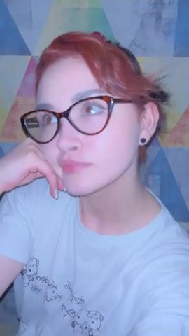 A young woman with red hair and glasses looks into the camera, touching her glasses. The background is a wall with a colorful geometric pattern.