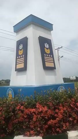The video shows a building with a blue and white facade, decorated with an emblem and what appears to be a rank insignia. The weather is overcast, and it seems to be raining lightly, with wet surfaces visible on the ground. The camera then pans to a paved road with a curb lined with black and white markings. The road leads into a landscape with scattered trees and greenery. In the distance, there are more buildings, streetlights, and utility poles with wires overhead. The overall impression is that of an institutional or governmental compound, possibly a military or police facility, given the insignia.