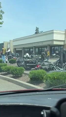 A group of people, likely motorcyclists, are gathered in a parking lot on a sunny day. Several motorcycles are parked in front of a building with a sign that reads "HIDA CO." Some of the individuals are wearing vests with patches, and some of the motorcycles have American flags attached. The scene appears to be set in a commercial area, possibly a strip mall, with a large tree providing shade. The overall mood seems casual and social, with people interacting and standing around their motorcycles. The time of day appears to be daytime, given the bright sunlight and clear sky. The location is Spanaway, United States of America.