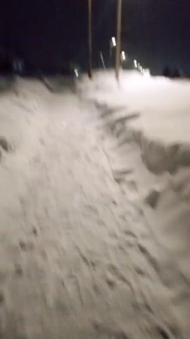 The video shows a snow-covered scene at night. A snow path is visible in the foreground, with a higher snowdrift on the right side of the path. Streetlights illuminate the scene, and snow is actively falling. There are utility poles along the street. Houses can be seen in the distance. Footprints are visible in the snow path.